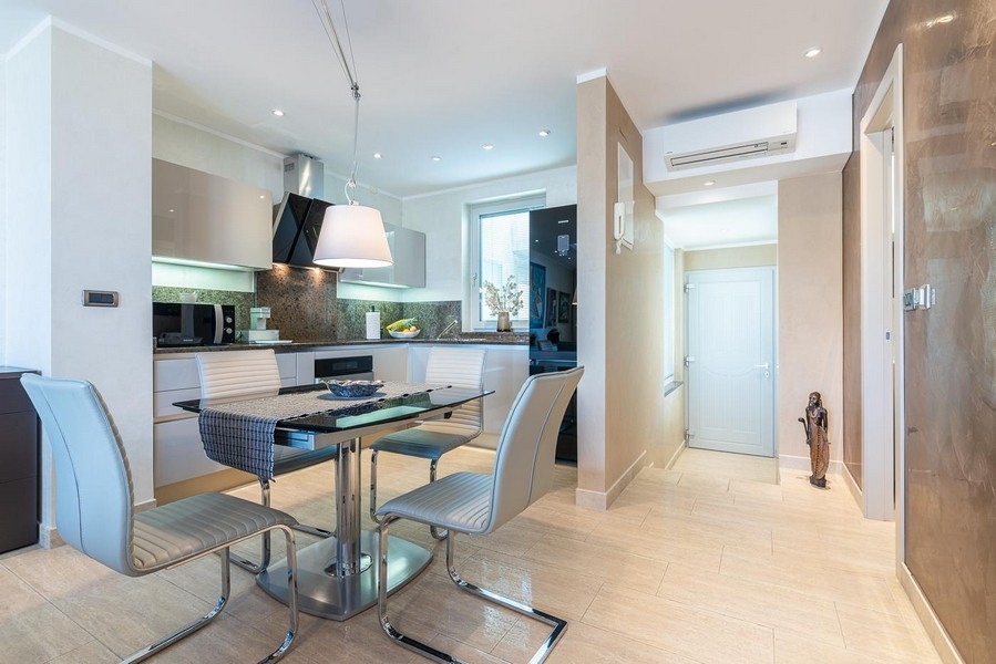 Moderní jídelní kout se skleněným stolem a výhledem do moderní kuchyně a klimatizovaného vstupního prostoru.