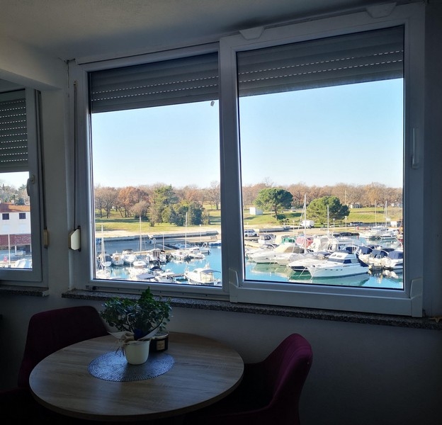 Útulný jídelní kout s kulatým stolem a výhledem na přístav přes velké okno v chorvatském apartmánu - Kupte si bydlení v Chorvatsku.