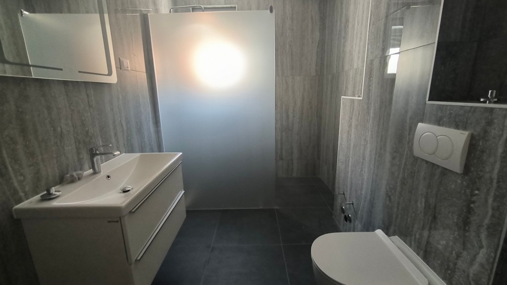 Moderně navržená koupelna se sprchovým koutem, nemovitost v Chorvatsku v Šimuni, zdůrazněná šedými dlaždicemi a jasným světlem.