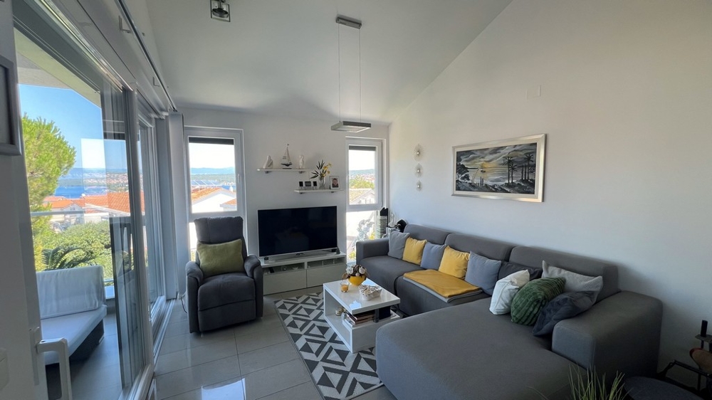 Moderní obývací pokoj s velkou TV, šedou pohovkou a výhledem na moře přes velká okna.