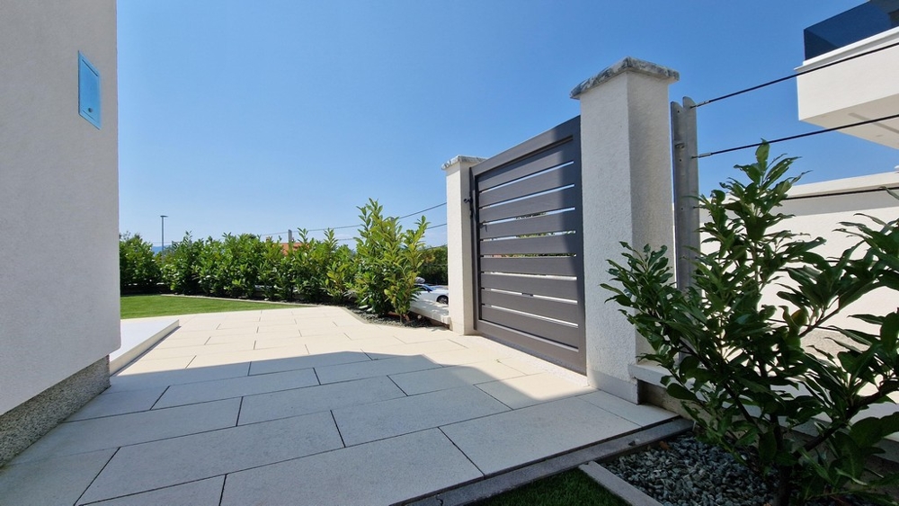 Vstupní brána do moderní budovy s dlážděným nádvořím a zelení.