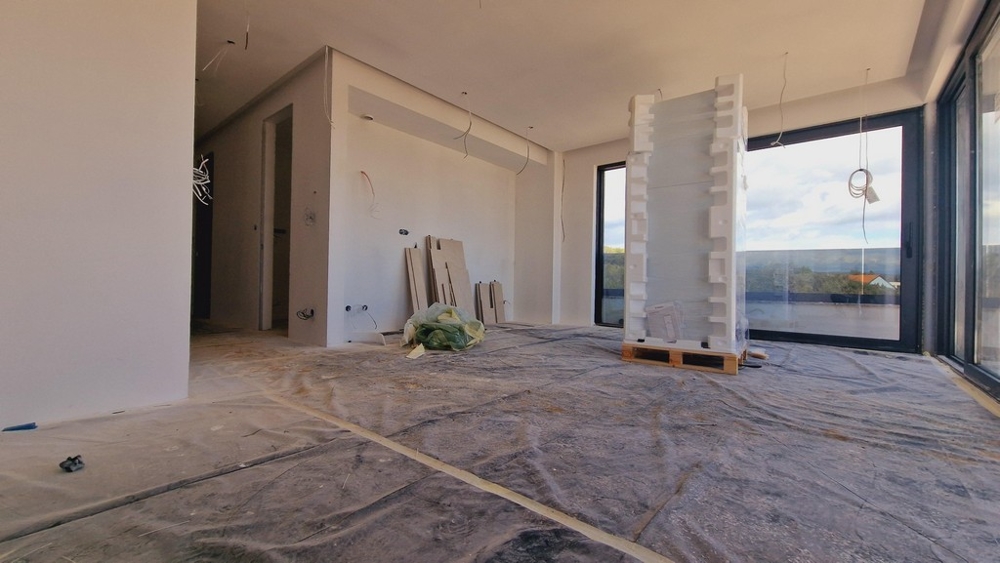 Obytná plocha novostavby ve fázi dokončování, s výhledem na velká prosklená okna a okolní přírodu.