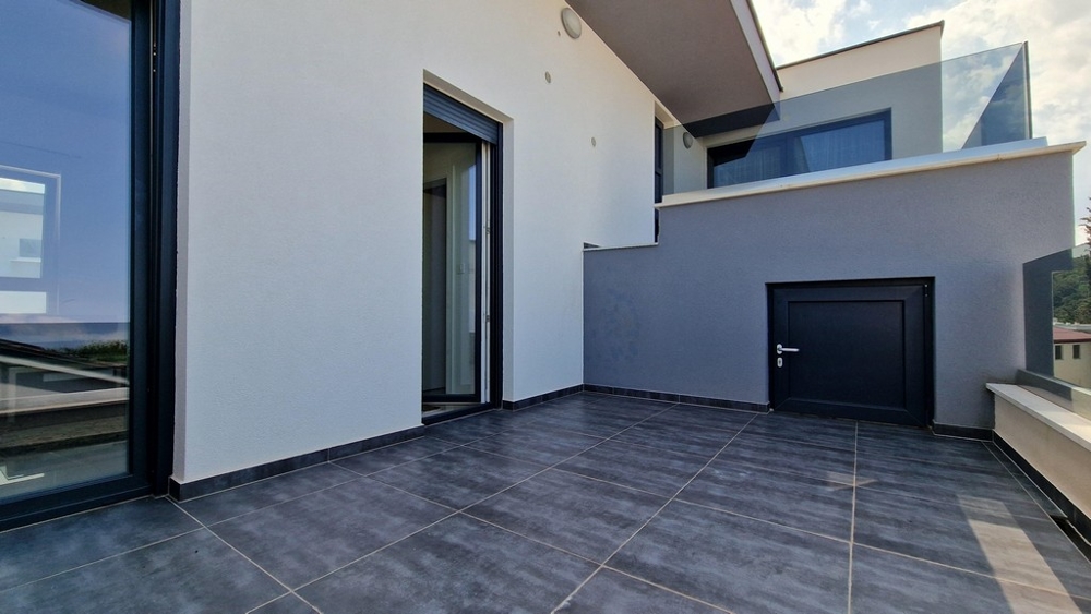 Moderní terasa s šedou dlažbou, proskleným zábradlím a vstupem do bytu.