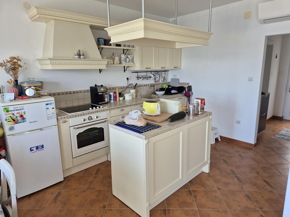 Světlá, otevřená kuchyně s béžovými skříňkami, ostrůvkem na vaření a dlážděnými podlahami.