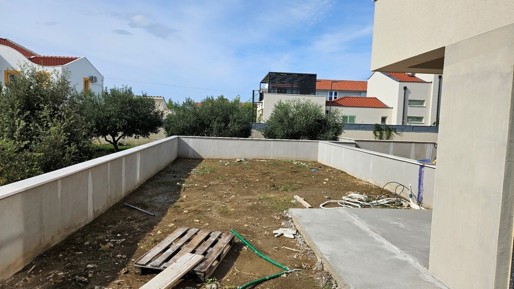 Venkovní plocha s nezpevněnou zemí, nízkou zídkou a několika stromy v pozadí.