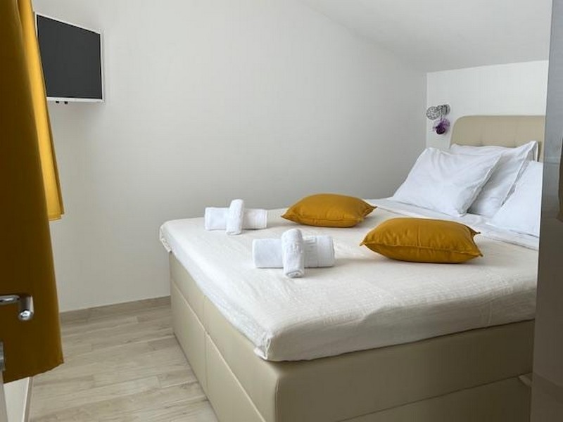 Ložnice s velkou postelí, žlutými polštáři a televizí na zdi.