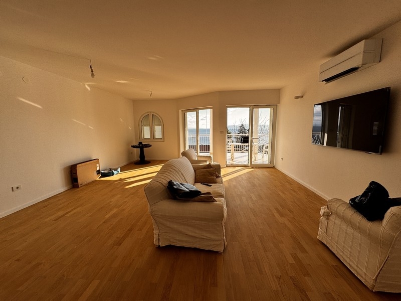 Světlý obývací pokoj s bílou pohovkou, nástěnnou TV a panoramatickými okny s výhledem na moře