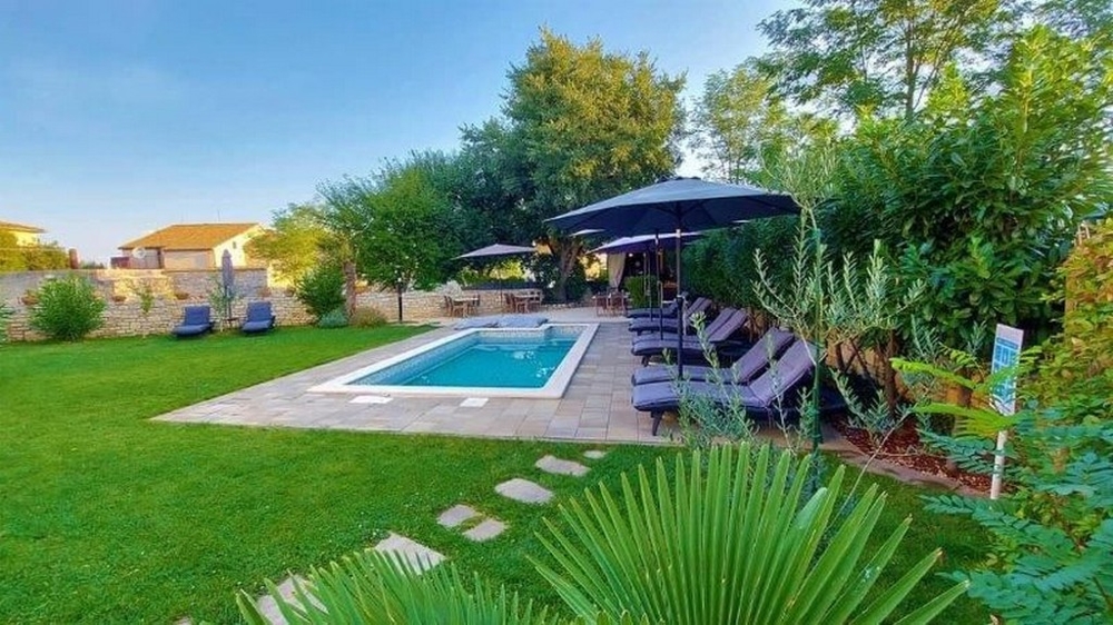 Pohled do zahrady na tříbytový dům na prodej na Istrii s bazénem, ​​lehátky, slunečníky a bujnou zelení.