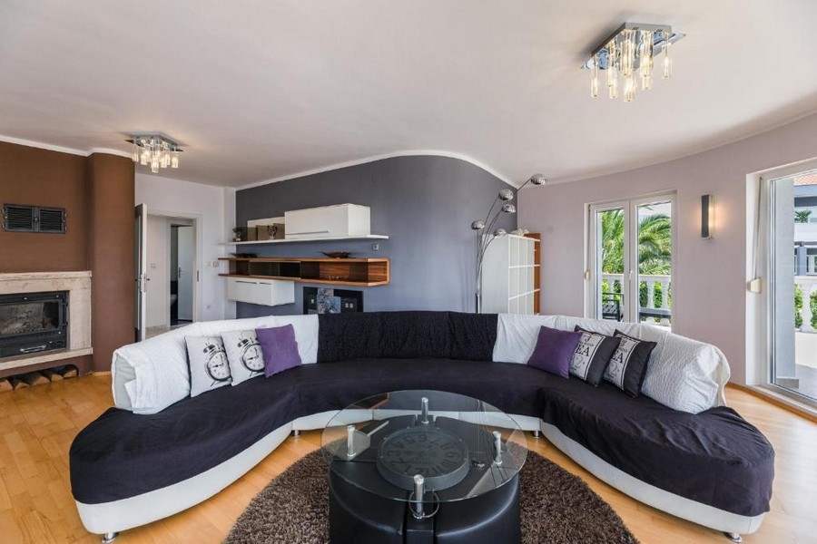 Elegantní obývací pokoj v chorvatském činžovním domě s výhledem na moře, s velkou zakřivenou pohovkou a kulatým skleněným stolem na hnědém koberci, krbem a otevřeným balkonem.