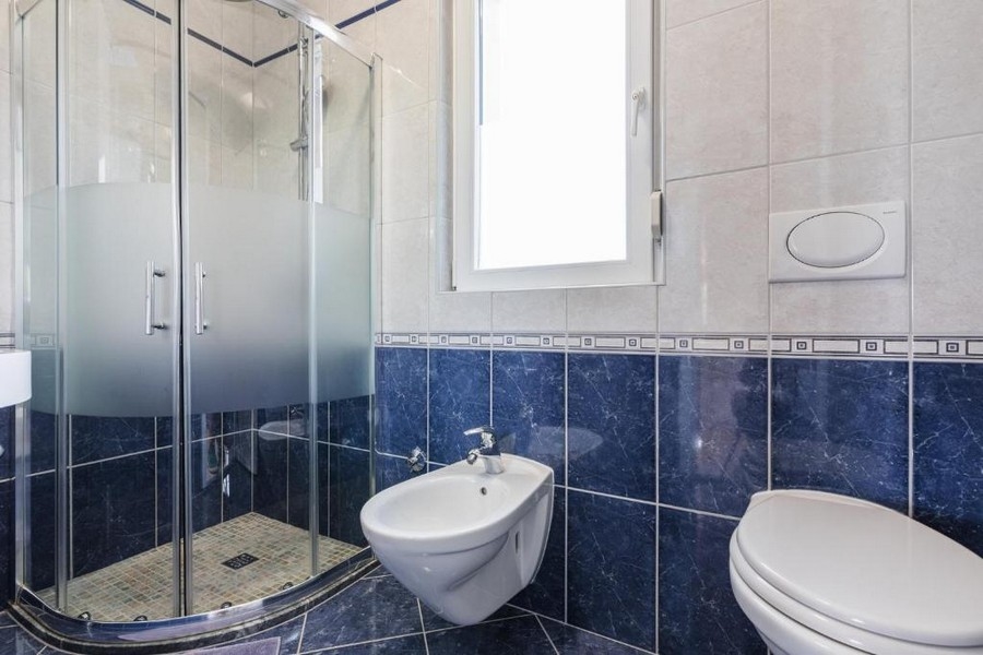 Koupelna v nemovitosti v Chorvatsku, bytový dům s výhledem na moře, s rohovou sprchou, bidetem a toaletou, světlé stěny a tmavě modré dlaždice