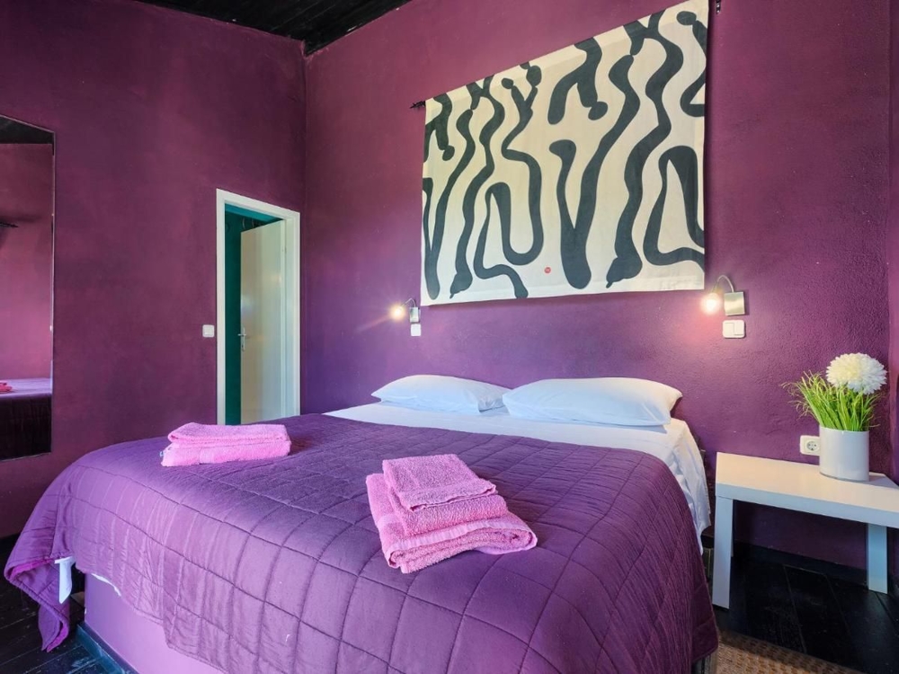 Ložnice v nemovitosti v Chorvatsku, s fialovými stěnami, velkým obrazem s abstraktními vzory, dvěma nočními lampami a lůžkovinami v odpovídajícím fialovém odstínu