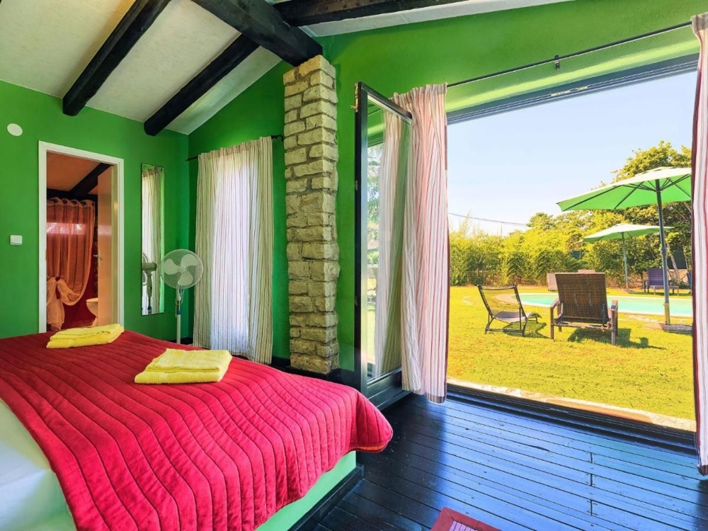 Světlá ložnice s výhledem do zahrady, součást nemovitosti v Chorvatsku, se zelenými stěnami, červeným přehozem a přístupem na terasu s lehátky a slunečníkem