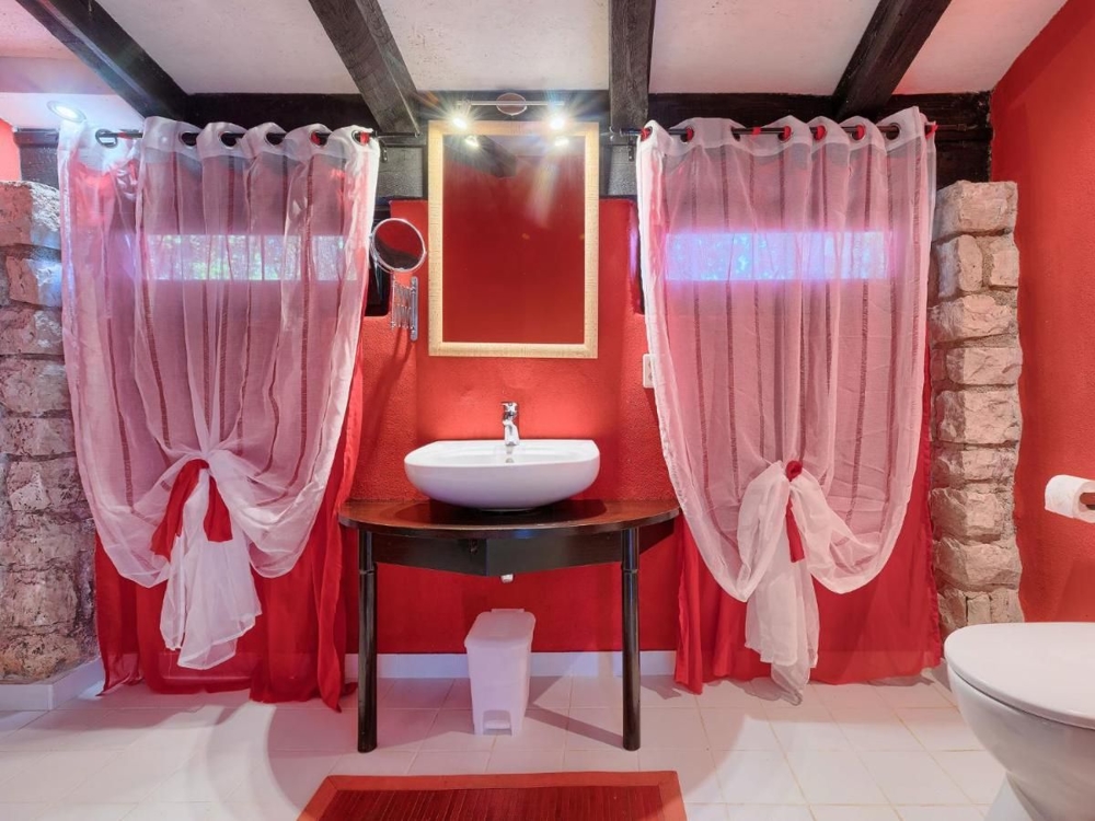 Koupelna nemovitosti v Chorvatsku s červenými stěnami, bílým vodovodem, umyvadlem na rustikálním stole a průhlednými červenými závěsy na oknech
