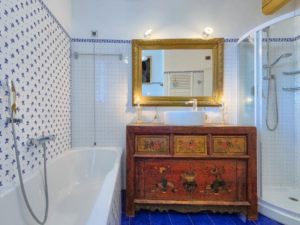 Koupelna v chorvatské nemovitosti zdobená modrobílými dlaždicemi, vanou, sprchovým koutem a starožitnou barevnou skříňkou pod zlatým zrcadlem