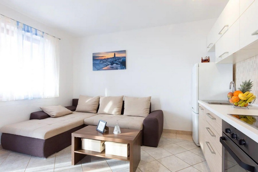 Útulný obývací pokoj s rohovou pohovkou, konferenčním stolkem a výhledem do kuchyně, zařízený v jemných barvách a obrázkem moře.