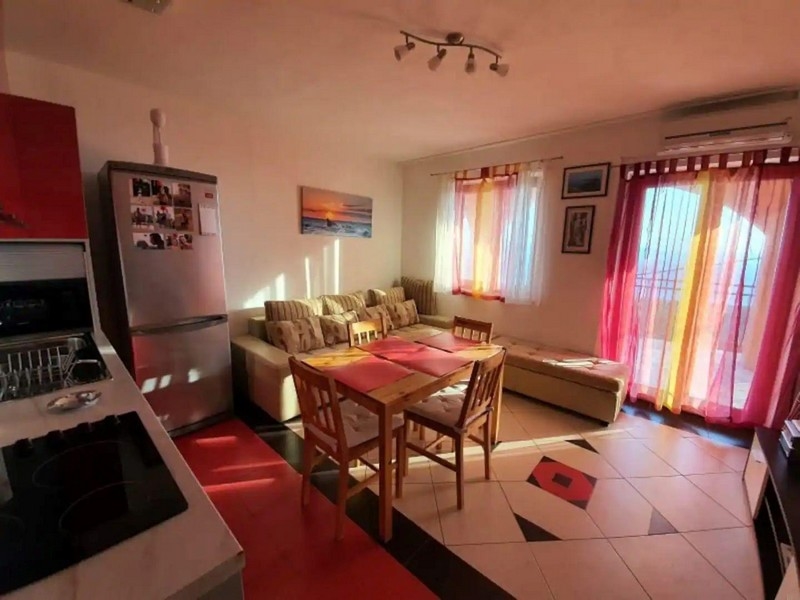 Prosluněný obývací a jídelní kout s červenými a žlutými akcenty, vybavený jídelním stolem, pohovkou a přístupem na balkon.