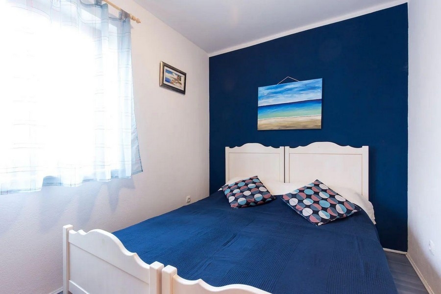 Ložnice s manželskou postelí, modrou akcentovou stěnou a námořní dekorací, osvětlená přirozeným světlem z okna.
