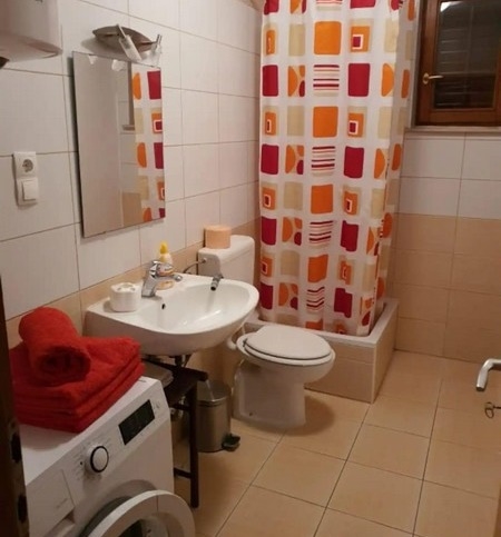 Funkční koupelna se sprchovým závěsem, pračkou a červenými a oranžovými doplňky, vybavená umyvadlem, toaletou a malým oknem.