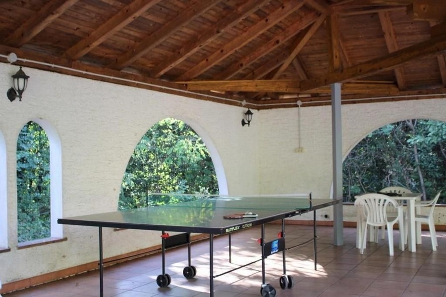 Krytá venkovní plocha se stolem na stolní tenis uprostřed, bílými plastovými židlemi a dřevěnou střechou. Stěny mají klenuté okenní otvory s výhledem do zeleně.