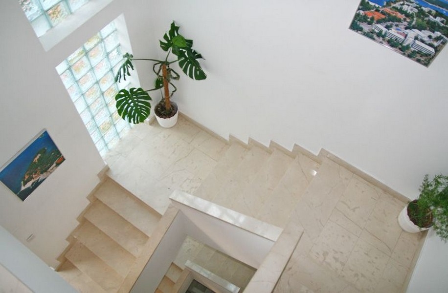 Schodiště se světlými mramorovými schody, velká okna se skleněnými tvárnicemi, zelené rostliny v bílých květináčích a obrazy visící na stěnách.