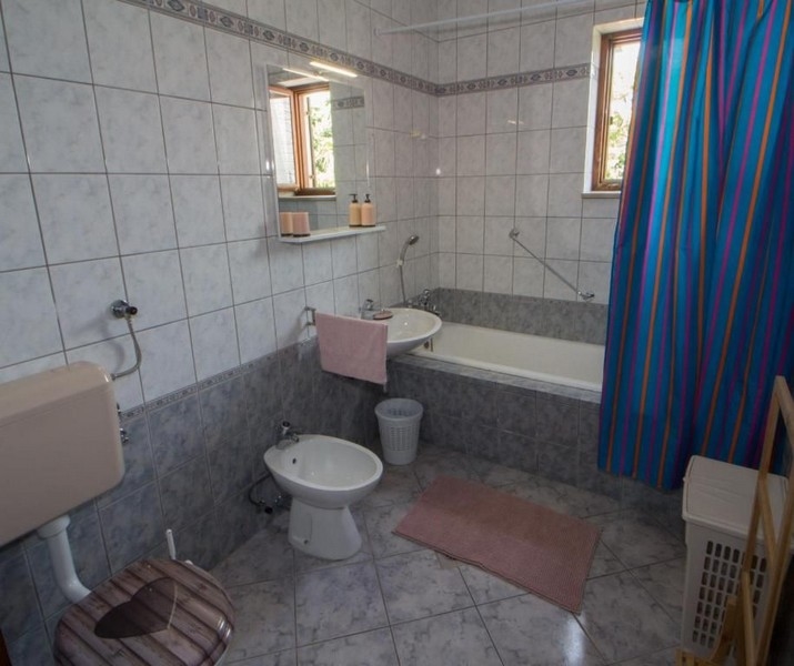 Koupelna s bílými a šedými obklady, vanou, umyvadlem, bidetem, toaletou a modrým sprchovým závěsem. Malá okna propouštějí přirozené světlo.