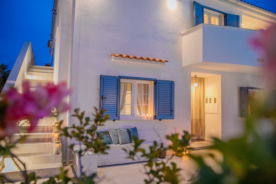 Čelní pohled na středomořský dům s modrými okenicemi a venkovním posezením za soumraku, osvětlené měkkým světlem
