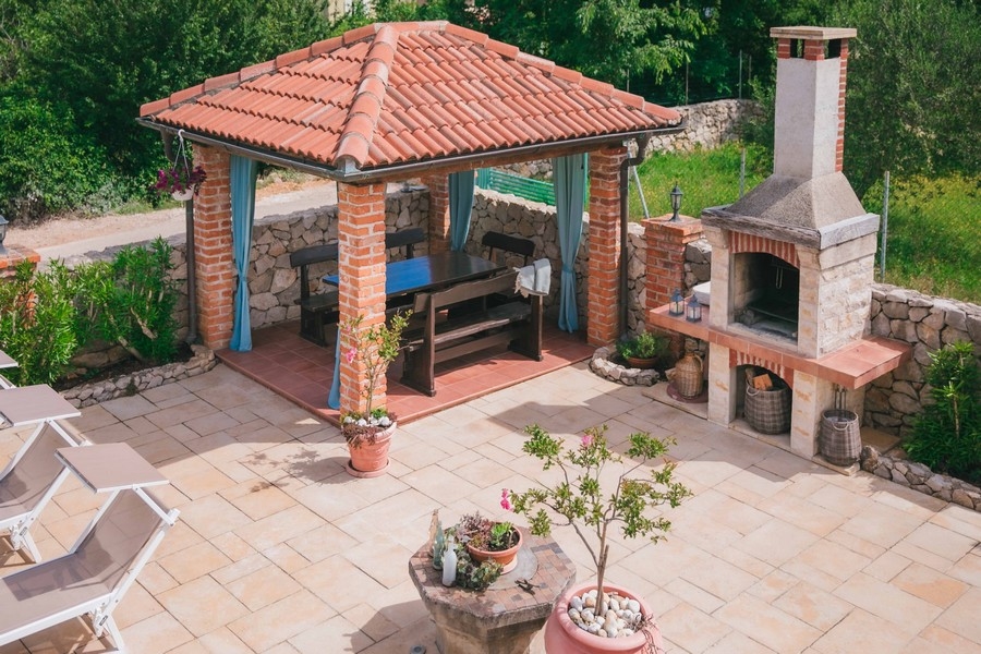Venkovní gril se zděným altánem, dřevěným jídelním stolem a kamennou pecí na dlážděné terase