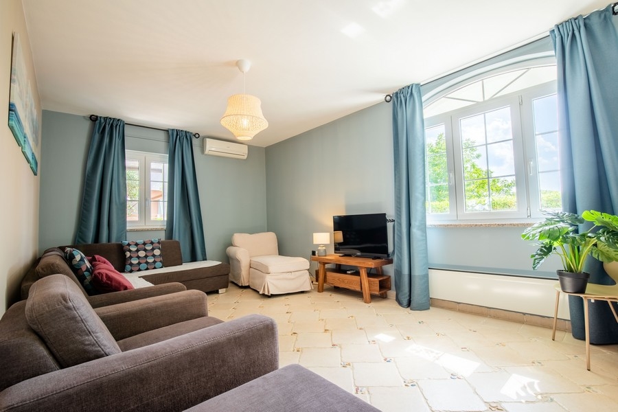 Útulný obývací pokoj s několika pohovkami, TV a velkými okny s modrými závěsy, které propouštějí přirozené světlo