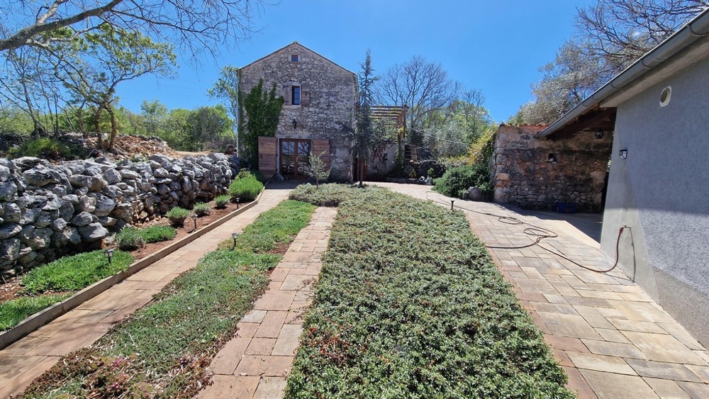 Udržovaná zahrada s dlážděnou cestou a kamenným domem v pozadí.