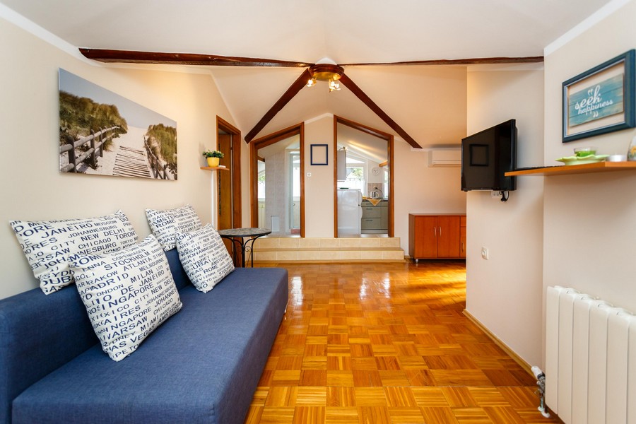 Obývací pokoj se sedací soupravou, dřevěnou podlahou a trámovým stropem vede do malého kuchyňského koutu