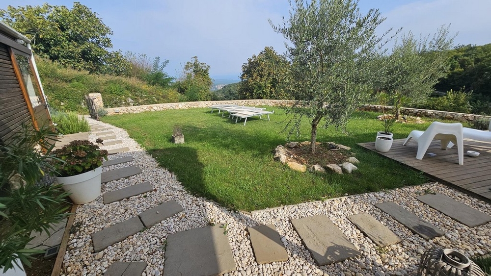 Udržovaná zahrada s olivovníkem a lehátky, obklopená přírodou a kamennou zdí.