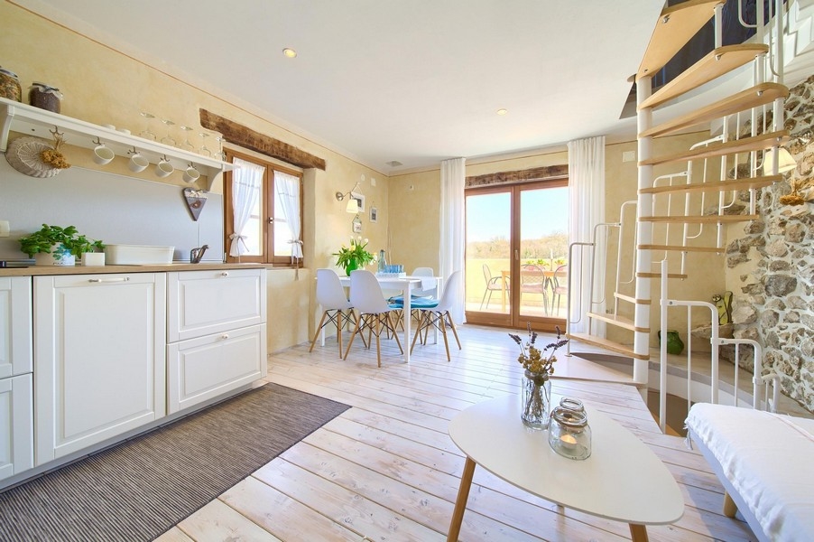 Otevřený obývací prostor s moderní kuchyní, jídelním stolem a přístupem na terasu, zařízený světlým nábytkem.