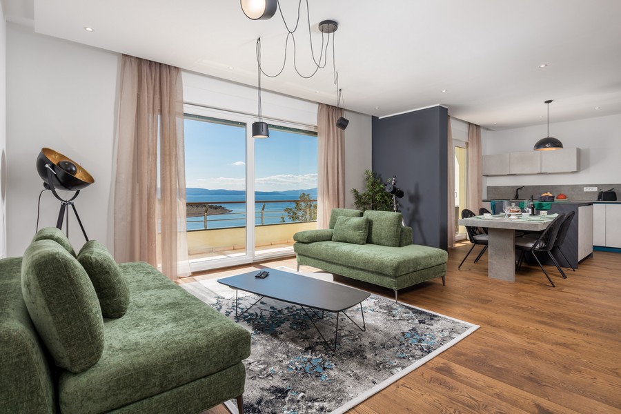 Obývací pokoj s velkými okny, stylovým nábytkem a výhledem na moře, v pozadí otevřená kuchyně.