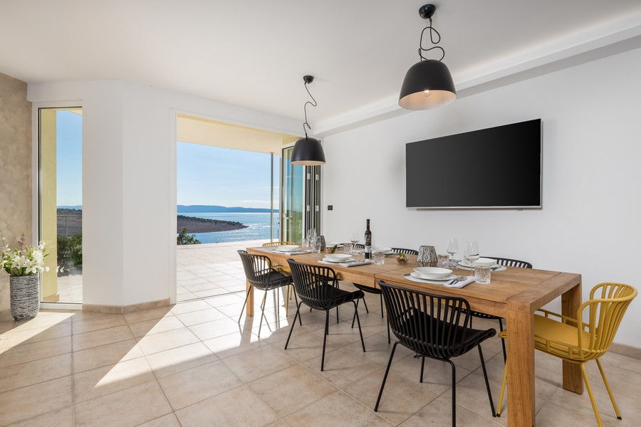 Jídelna s velkým dřevěným stolem, moderními židlemi a přímým přístupem na terasu s výhledem na moře.