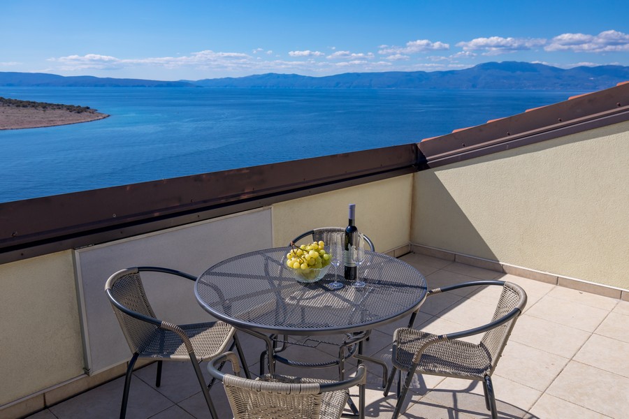 Terasa se stolem a židlemi, výhled na moře a okolní kopce v pozadí.