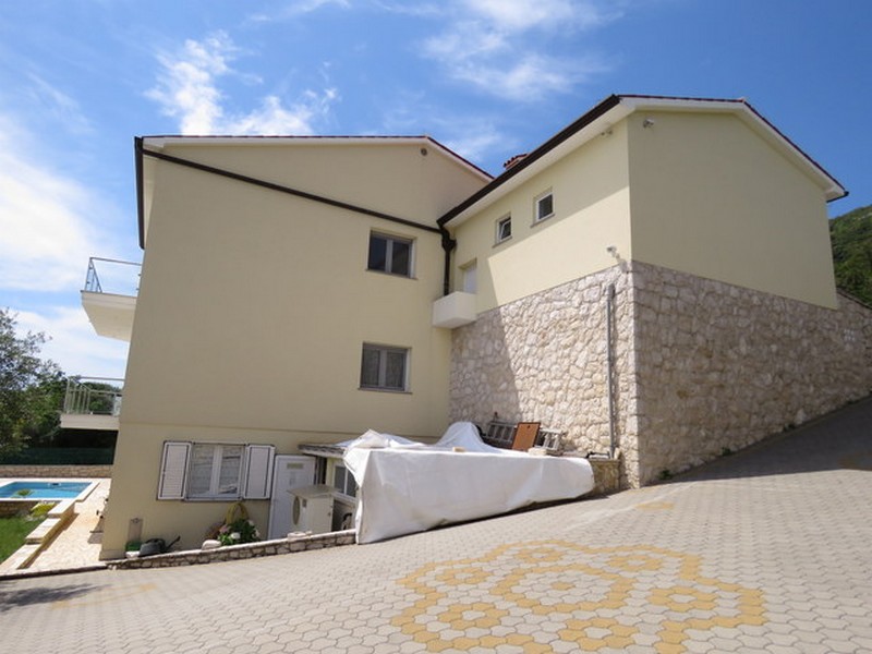 Vnější pohled na dům s dlážděným parkovištěm a bazénem v pozadí.